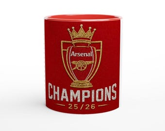 May include: A red and white ceramic mug featuring the Arsenal logo, a gold crown, and the word "CHAMPIONS" in white. The mug has a red interior and the text "25/26" below the word "CHAMPIONS".
