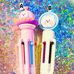 May include: Two multi-colored pens with cartoon bear characters on the top. The pen on the left has a pink and purple body with a pink and purple spaceship on the top. The pen on the right has a blue and white body with a blue and white bear on the top.