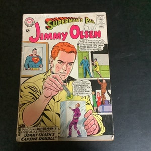 May include: A comic book cover featuring Superman's Pal Jimmy Olsen. The cover art shows Jimmy Olsen holding a device and looking at a glass case with a figure inside. The text on the cover reads "Superman's Pal Jimmy Olsen" and "Featuring Superman's Look-Alike Friends... Jimmy Olsen's Captive Double!"