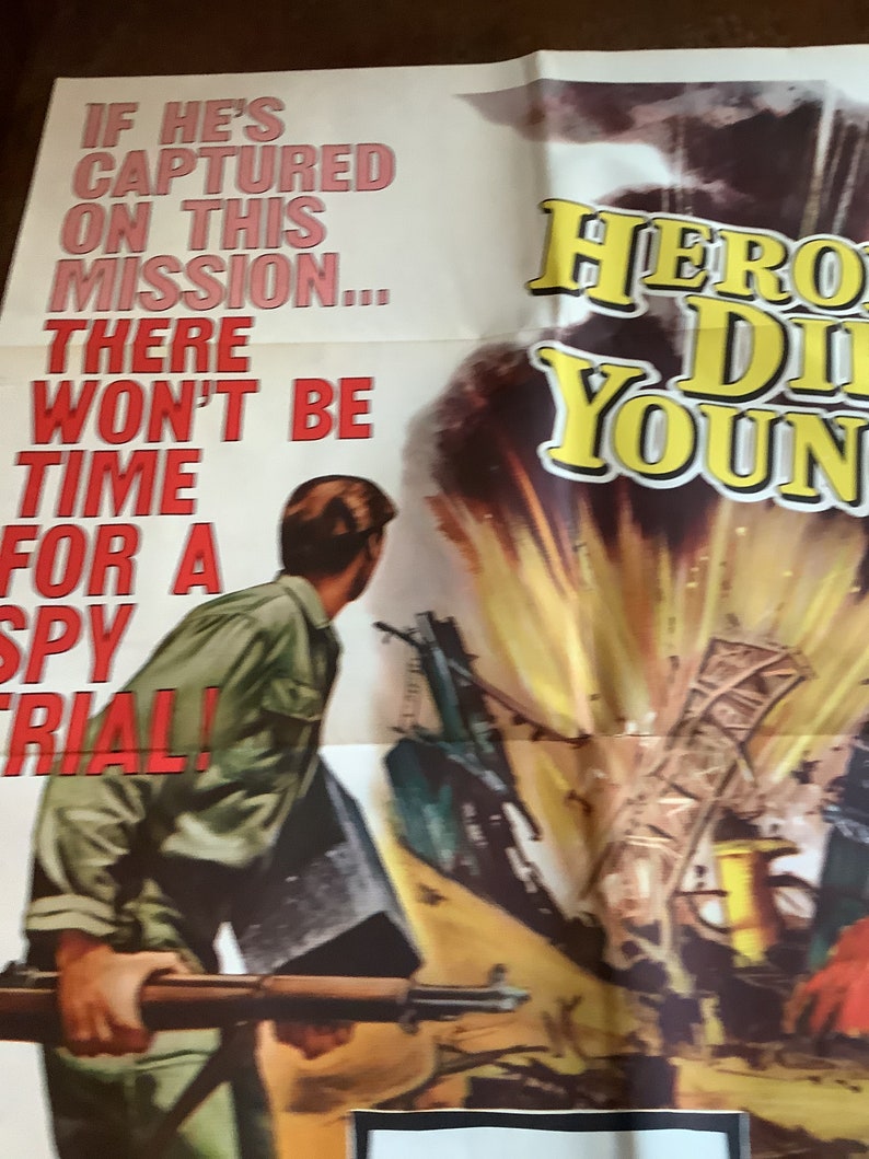 Op de afbeelding: Een vintage filmposter voor de film "Hero Die Young" met een man in een groen shirt die een geweer vasthoudt. De poster heeft een geel en rood kleurenschema met de titel in grote gele letters. De tekst op de poster luidt: "Als hij tijdens deze missie wordt gevangen genomen... is er geen tijd voor een spionageproces!"