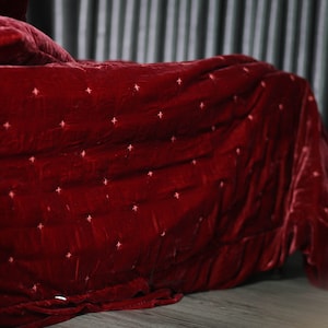 May include: A red velvet quilted blanket with a subtle diamond pattern. The blanket is folded and draped over a bed.