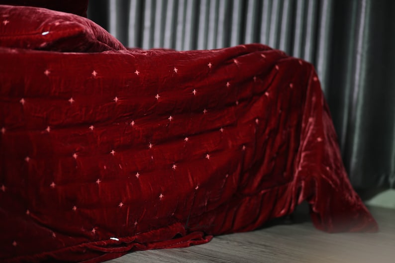 May include: A red velvet quilted blanket with a subtle diamond pattern. The blanket is folded and draped over a bed.