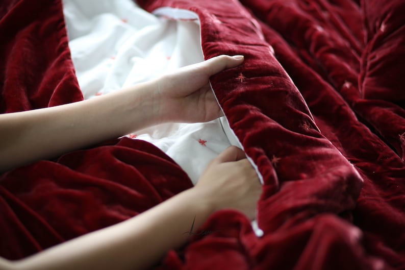 May include: A person is holding a red velvet blanket with white stars embroidered on it. The blanket is folded in half and the person is holding the edge of the blanket with their right hand.