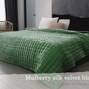Può includere: Una coperta in velluto di seta di gelso trapuntata di colore verde su un letto con cuscini bianchi e neri. La coperta è cucita a mano da artigiani. "Mulberry silk velvet blanket" e "HAND - STITCH BY ARTISANS" sono scritti in bianco su sfondo nero sotto la coperta.