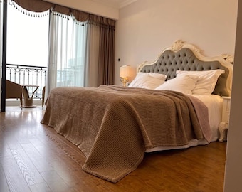 Op de afbeelding: Een bruine wafeldekbed op een bed met een witte hoofdeinde en witte beddengoed. Het bed staat in een slaapkamer met een balkon en houten vloer.