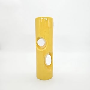 May include: A tall, cylindrical yellow ceramic vase with two large, circular cutouts. The vase has a glossy finish and stands upright against a white background. The vase is a decorative item.