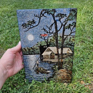 May include: A rectangular plaque featuring a textured, glittery landscape scene of a stilt house over water at night. The image depicts a small boat with a person, a full moon, and tall trees against a dark blue and black background with gold accents.