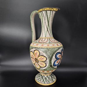 May include: A ceramic pitcher featuring a speckled cream base with hand-painted floral motifs in yellow, blue, and brown. The vessel has a tall neck, a curved handle, and a flared rim, decorated with geometric green and yellow patterns.