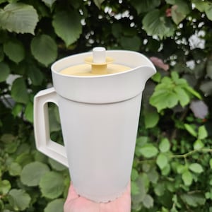 Può includere: Una caraffa in plastica bianca dotata di manico laterale e coperchio giallo chiaro con un piccolo pomello centrale bianco. La caraffa presenta un corpo affusolato con un beccuccio sul bordo superiore, progettata per servire liquidi.