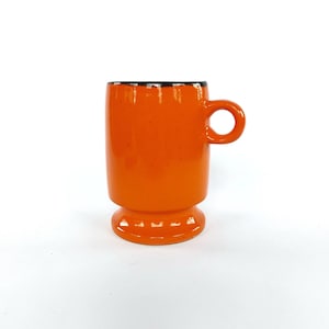 Könnte beinhalten: Orangefarbene Keramiktasse mit schwarzem Rand und einem ausgestellten Boden. Die Tasse hat einen einfachen, abgerundeten Griff.