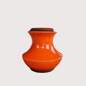 May include: A vibrant orange ceramic vase with a glossy finish. The vase has a unique shape, wider at the base and top, with a narrower midsection. The rim is a darker shade of brown. A decorative piece for home decor.