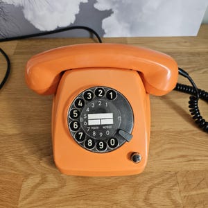 Può includere: Un telefono a disco di colore arancione brillante con un quadrante circolare nero che presenta numeri bianchi da uno a zero. Il centro del quadrante include due etichette rettangolari bianche e il testo in tedesco FEUER e NOTRUF. È collegato un cavo nero a spirale.