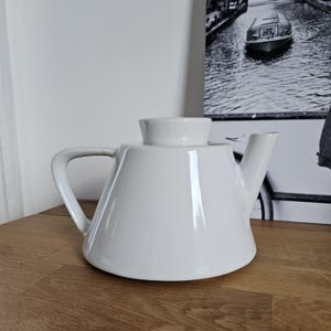 Könnte beinhalten: Eine glänzend weiße Keramik-Teekanne mit breitem, konischem Boden, einem geschwungenen Griff auf der linken Seite, einem kurzen, abgewinkelten Ausguss auf der rechten Seite und einem passenden, mittig platzierten runden Deckel oben.