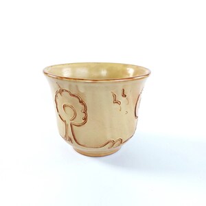 Pode incluir: Uma chávena de cerâmica de cor creme com um desenho de árvore castanho. A chávena é ligeiramente alargada na parte superior e tem um fundo arredondado.
