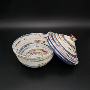 May include: A round bowl with a lid, crafted from rolled paper. The bowl is white with colourful paper edges. The lid is slightly ajar, showing the inside. The lid has a small red knob.