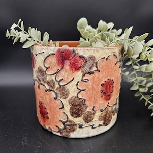 Puede incluir: Jardinera cilíndrica de cerámica con un diseño floral en tonos melocotón, rojo y marrón, delineado en negro. La jardinera está llena de una planta de hojas verdes. La jardinera tiene una base de color crema.