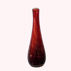 Può includere: Un vaso di vetro rosso, alto e sottile, con un collo lungo e una base arrotondata. Il vaso ha una superficie strutturata con piccoli accenti bianchi a pois. Il vaso è alto circa 30 cm.