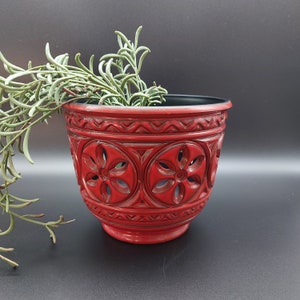 May include: A red ceramic planter with a decorative floral design. The planter has a black rim and is empty.