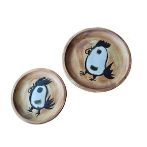 May include: Two round, brown ceramic plates with a whimsical bird design. The birds have white bodies, black wings and feet, and a black crest. The plates have a rustic, hand-painted appearance.