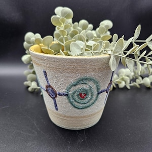 Op de afbeelding: Een kleine, beige keramische plantenbak met een getextureerd oppervlak en een gele binnenkant. De plantenbak is versierd met blauwe en groene spiraalontwerpen en bevat een lichtgroene plant. De plantenbak is ongeveer 10 cm hoog.