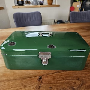 May include: A vintage green metal bread box with a hinged lid and a silver clasp. The box has a handle on top and some areas of wear and tear, giving it a rustic appearance. The bread box is sitting on a wooden table.