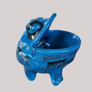 Puede incluir: Un cuenco de cerámica con forma de pelícano en un vibrante esmalte azul. El ave con la boca abierta tiene una superficie texturizada con detalles en negro y dorado. El cuenco está sostenido por tres pequeñas patas.