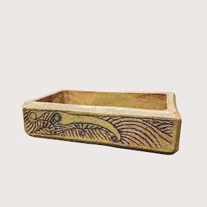 May include: A rectangular ceramic container with a light yellow-brown glaze. The sides are decorated with a carved design featuring flowing lines and a stylized organic shape. The container has a slightly rough, textured appearance.