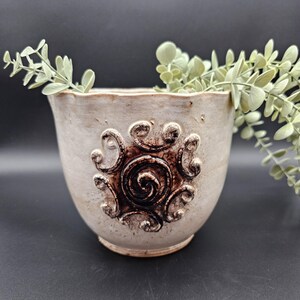 May include: A cream-colored ceramic planter with a decorative brown spiral and swirl design. The planter has a textured surface and a slightly ruffled rim. Greenery is placed inside the planter.