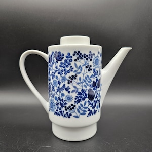 Könnte beinhalten: Weiße Keramik-Teekanne mit blauem Blumenmuster. Die Teekanne hat einen gebogenen Griff, einen Ausguss und einen Deckel. Das blaue Muster umfasst Blumen, Beeren und Blätter. Ein klassisches Design zum Servieren von Tee.