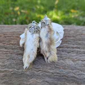 Könnte beinhalten: Zwei Hasenpfoten-Schlüsselanhänger. Jeder Schlüsselanhänger hat eine echte Hasenpfote mit hellbraunem und beigem Fell, die von einer dekorativen, silberfarbenen Kappe gekrönt wird. Die Schlüsselanhänger werden auf einer Holzoberfläche mit verschwommenem grünem Hintergrund gezeigt.