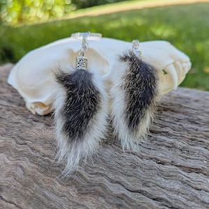 May include: A pair of dangle earrings featuring fluffy, black and white fur pendants. The earrings hang from ornate silver-colored metal findings. The earrings are displayed on a white skull against a blurred green background.