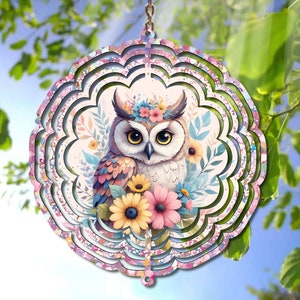 May include: A pink and white metal wind spinner with a colorful owl and flowers. The owl has a flower crown and is surrounded by pink, yellow, and blue flowers.