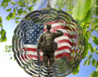 May include: A decorative wind spinner featuring a soldier saluting in front of an American flag. The design incorporates a camouflage pattern around the edges, with a chain for hanging. The background shows a blue sky and green foliage.