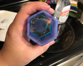 May include: A purple hexagonal resin mold with a clear center and green glitter. The mold is being held in a hand.