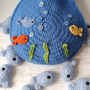 May include: A blue crocheted bag with a handle, decorated with crocheted fish in orange, yellow, and blue, along with green seaweed and blue buttons. Several small blue crocheted fish are scattered around the bag.