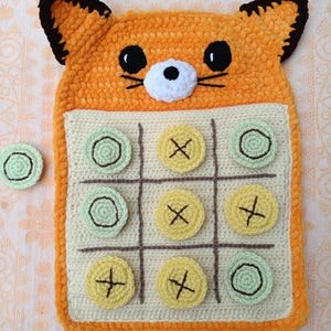 Può includere: Un gioco del tris all'uncinetto a forma di volpe nei colori arancione, bianco e marrone. La tavola presenta una griglia con nove quadrati, inclusi pezzi circolari gialli con ricamo a X marrone e pezzi verde chiaro con ricamo a O marrone.