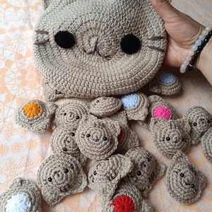 Peut inclure: Une grande pochette crochetée beige en forme de chat avec des yeux noirs et des moustaches brodées, entourée de nombreuses petites appliques crochetées en forme de chat, chacune avec un petit centre circulaire blanc, rouge, orange, bleu ou rose.