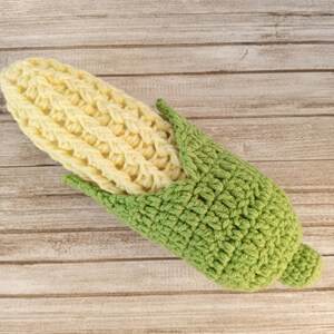 Könnte beinhalten: Ein gehäkeltes Mais-Spielzeug mit gelbem Kolben und grünen Blättern. Das Spielzeug ist aus Garn gefertigt und eine lustige und fantasievolle Ergänzung für jedes Spielzimmer.
