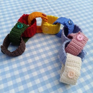 May include: A chain of interconnected crocheted fabric loops in various colors including brown, dark green, red, orange, yellow, blue, purple, pink, and white. Each loop is secured with a matching plastic button fastened through the fabric.