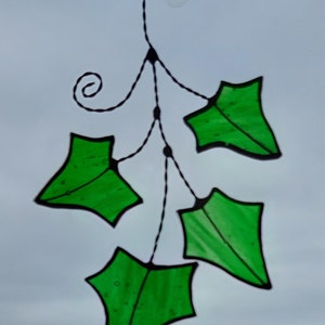 May include: A stained glass window hanging with a black wire frame and five green leaves. The leaves are shaped like a traditional holly leaf.
