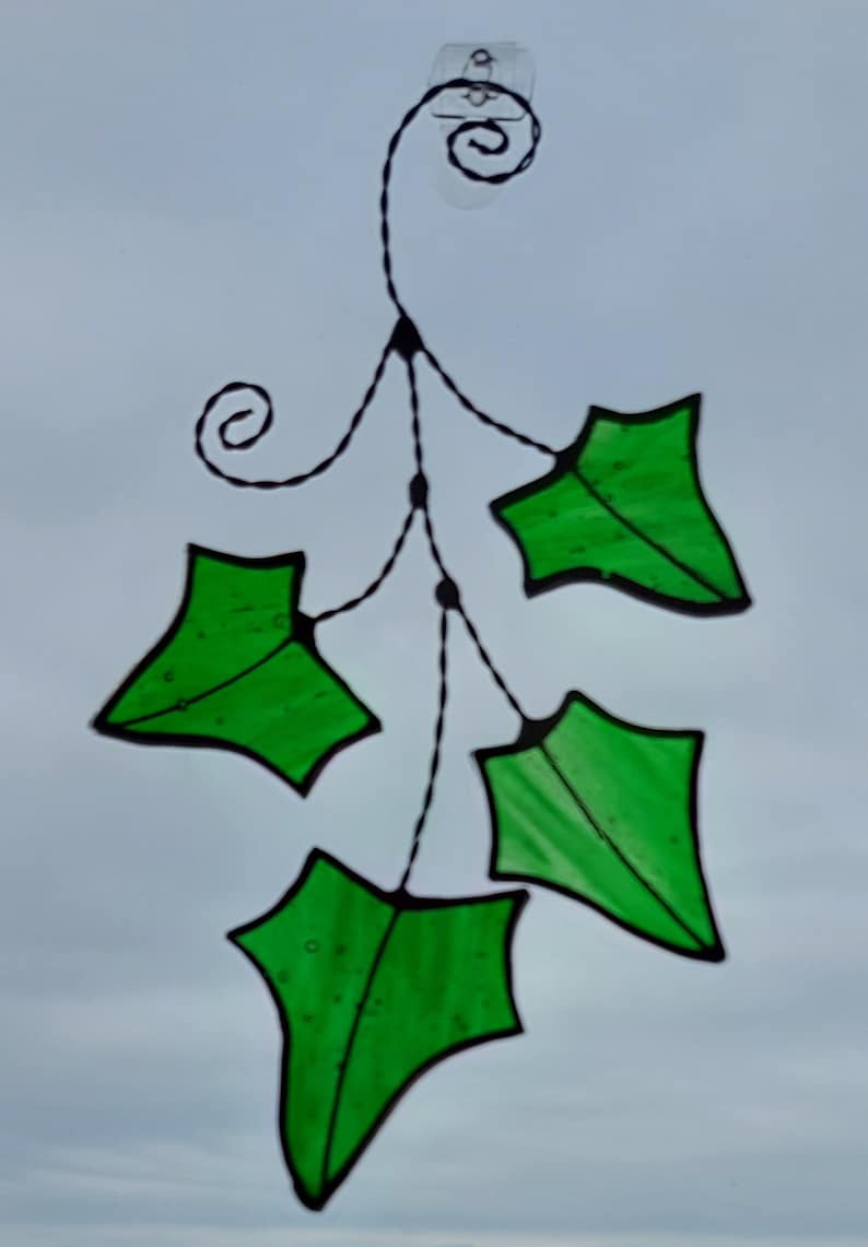 May include: A stained glass window hanging with a black wire frame and five green leaves. The leaves are shaped like a traditional holly leaf.