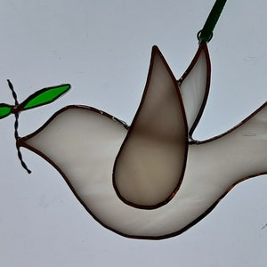 May include: Stained glass dove with a green olive branch in its beak. The dove is white with brown outlines and a green ribbon for hanging. A symbol of peace and hope.