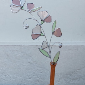 May include: A decorative stained glass flower arrangement in a brown vase. The flower has pink and white petals with green leaves. The vase is sitting on a brown wicker surface.