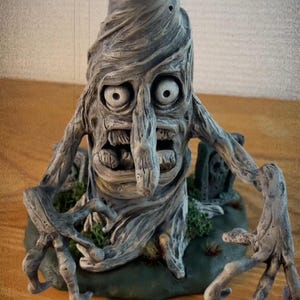 May include: A gray, twisted tree sculpture with a grotesque face, large eyes, and a long nose. The tree is sitting on a green base with a small graveyard in the background.
