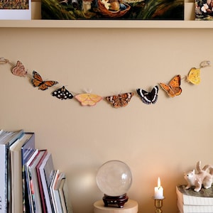 May include: A decorative garland featuring a series of eight distinct butterfly and moth illustrations in various colors including orange, yellow, brown, and black, strung together on a thin twine cord against a plain light beige wall surface.