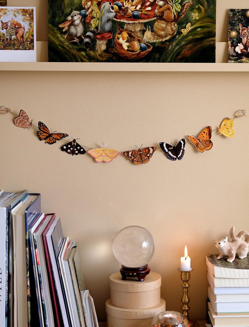 May include: A decorative garland featuring a series of eight distinct butterfly and moth illustrations in various colors including orange, yellow, brown, and black, strung together on a thin twine cord against a plain light beige wall surface.