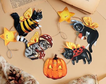 May include: A decorative garland featuring flat, illustrated cutouts of black and grey cats with yellow butterfly wings, orange stars, and a central orange jack-o'-lantern pumpkin, all connected by thin brown twine string.