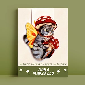 Peut inclure: Un chat gris avec une aile de papillon jaune et un chapeau de champignon rouge. Le chat tient un champignon rouge dans ses pattes. L'image est un marque-page magnétique avec le texte "Doro Marcello" et "Magnetic Bookmark - Signet Magnétique".