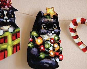 May include: A decorative garland featuring a black and white cat illustration wrapped in colorful Christmas lights with a gold star on its head, positioned between a cat in a gift box and a red and white striped candy cane shape on twine.