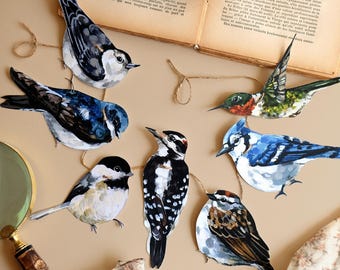 May include: A collection of six bird-shaped paper cutouts featuring a nuthatch, a swallow, a hummingbird, a chickadee, a woodpecker, a blue jay, and a sparrow, all connected by a thin brown twine string against a plain light tan surface.
